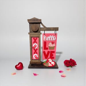 Puede incluir: Un letrero de madera del Día de San Valentín con las palabras "hello LOVE" y "XOXO" en rojo y blanco. Un corazón rojo y un par de fresas con un lazo están en la base. También hay dos rosas rojas y pétalos.