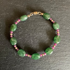 Puede incluir: Un brazalete de piedras preciosas verdes y rosas con cierre magnético. El brazalete está hecho con piedras verdes de forma ovalada y pequeñas cuentas rosas.