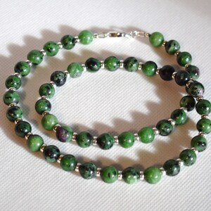 May include: A green and silver necklace made of round beads. The beads are a mix of green and black with silver accents. The necklace is made of natural stone and is approximately 18 inches long.
