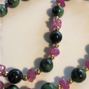 Puede incluir: Un collar de piedras preciosas verdes y negras con acentos de piedras preciosas rosas. El collar está hecho de cuentas redondas y está enhebrado en una cadena de oro.