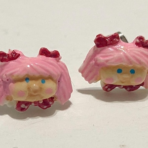 May include: A pair of pink and red plastic earrings shaped like a girl's face with blue eyes.