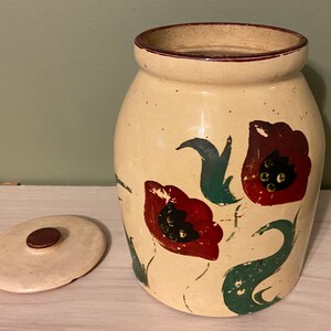 Könnte beinhalten: Eine antike Keramik-Keksdose mit cremefarbener Basis und braunem Deckel. Die Dose ist mit handbemalten roten und grünen Blumen verziert.