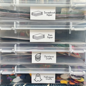 May include: Four clear plastic storage containers stacked on top of each other. Each container has a white label with black text and a simple icon. The labels read "Scrapbook Paper", "Plain Paper", "Christmas Crafts", and "Halloween Crafts".