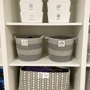 May include: White plastic storage bins labeled with black text and icons for pens, markers, bead trays, crayons, and pencils. Two gray and white striped rope baskets labeled "Imagination" and "Gaming". A gray and white polka dot fabric storage bin labeled "Barbie House".