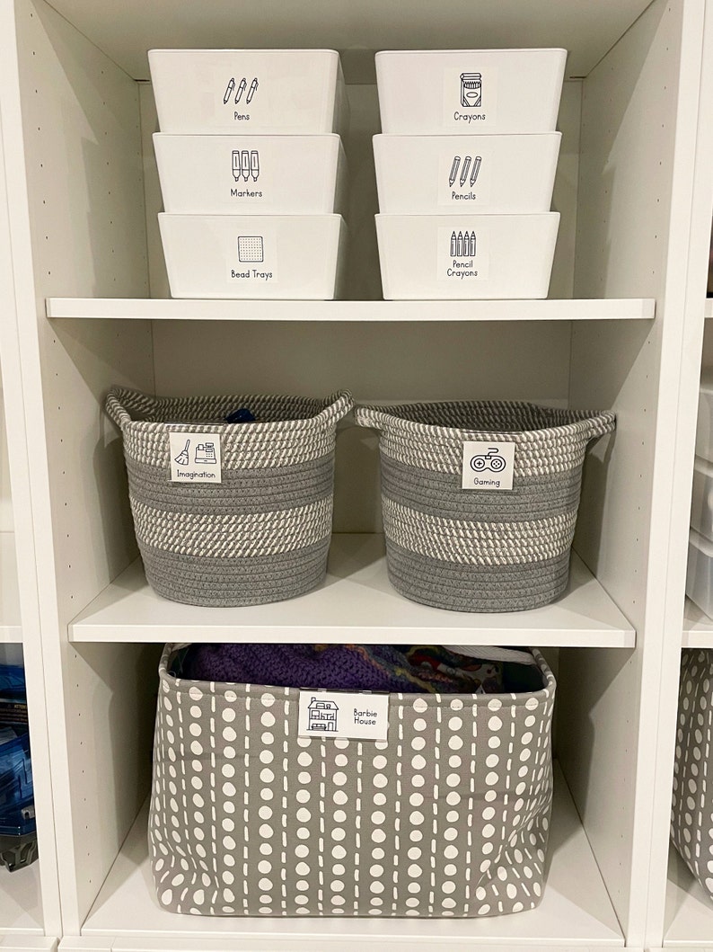 May include: White plastic storage bins labeled with black text and icons for pens, markers, bead trays, crayons, and pencils. Two gray and white striped rope baskets labeled "Imagination" and "Gaming". A gray and white polka dot fabric storage bin labeled "Barbie House".