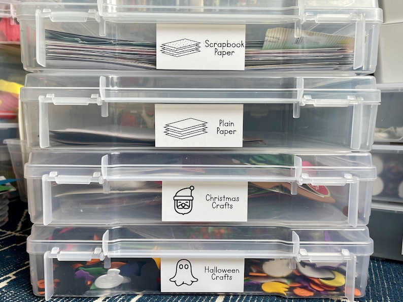 May include: Four clear plastic storage containers stacked on top of each other. Each container has a white label with black text and a simple icon. The labels read "Scrapbook Paper", "Plain Paper", "Christmas Crafts", and "Halloween Crafts".