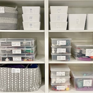 May include: White shelves with clear plastic storage bins and baskets labeled with black and white drawings and text. The labels include "Inspiration", "Crayons", "Pencils", "Markers", "Bead Trays", "Glitter", "Glue & Tape", "Construction Paper", "Christmas Crafts", "Halloween Crafts", "Ribbon Yarn & String", "Beads", "Sticker Beads", "Paint", "Kid's Crafts", "Foam Pieces", "Kinetic Sand".