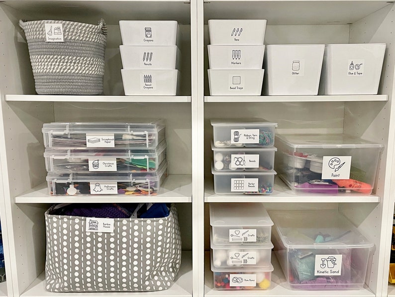 May include: White shelves with clear plastic storage bins and baskets labeled with black and white drawings and text. The labels include "Inspiration", "Crayons", "Pencils", "Markers", "Bead Trays", "Glitter", "Glue & Tape", "Construction Paper", "Christmas Crafts", "Halloween Crafts", "Ribbon Yarn & String", "Beads", "Sticker Beads", "Paint", "Kid's Crafts", "Foam Pieces", "Kinetic Sand".