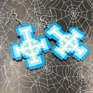 May include: A pair of pixelated cross earrings in shades of blue and white. The earrings are made of small, fused beads and have silver-colored ear wires. The background is a black surface with a white spiderweb design.