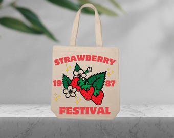 Puede incluir: Una bolsa de tela beige con un diseño gráfico rojo y blanco que dice "Strawberry Festival 1987". El gráfico presenta un grupo de fresas rojas con hojas verdes y flores blancas.