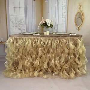 May include: A rectangular table with a champagne-colored ruffled skirt. The table is set with plates, silverware, and glasses. A floral arrangement and candles are in the center. The background features a window and a decorative mirror.