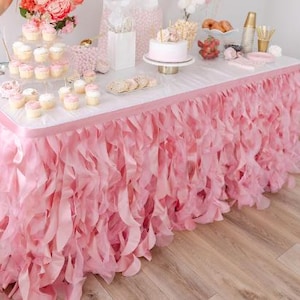 May include: Pink table skirt with ruffled edges. The skirt is made of tulle and is perfect for adding a touch of elegance to any party or event.