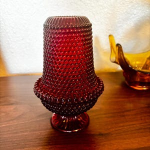 May include: A deep red glass vase featuring a textured hobnail pattern across its cylindrical body. The vase has a flared base with vertical ribbing and a rounded, wider midsection that tapers towards the top opening, which also has vertical ribbing.