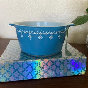 May include: A vintage turquoise Pyrex bowl with white snowflake and scalloped design. The bowl is set on a box with a holographic mermaid scale pattern. The bowl has two handles and a light blue interior.