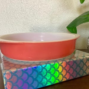 Puede incluir: Un molde para hornear Pyrex redondo, de color coral, con interior blanco. El plato está sobre una caja con un patrón holográfico de escamas de sirena. Una planta de interior en una maceta espejada está en el fondo.