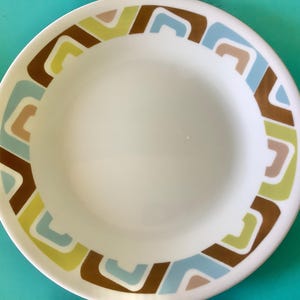 Puede incluir: Un plato redondo de cerámica blanca con un borde ancho decorado con un patrón geométrico repetitivo de formas en U entrelazadas en tonos marrón, azul claro, beige y verde lima, rodeando un centro circular blanco liso.
