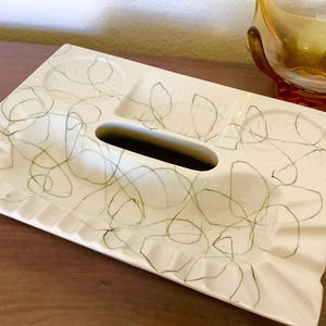 May include: A rectangular off-white ceramic tray featuring a central elongated oval opening and multiple recessed circular and rectangular compartments. The surface is decorated with a thin, dark green continuous line pattern.