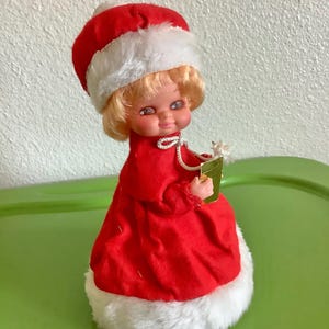 May include: A vintage Christmas doll wearing a red dress and white fur trimmed hat, holding a gold gift box.