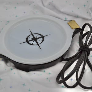 May include: A white and black electric serving dish with a compass design. The dish has a black base, gold-colored accents, and a black power cord. The dish is approximately 12 inches in diameter.