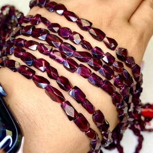 May include: A strand of dark red faceted gemstone beads. The beads are rectangular in shape and have a glossy finish.
