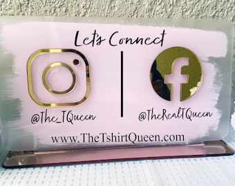 May include: A clear acrylic sign with a pink and white painted background. Gold Instagram and Facebook logos are displayed with the text "Let's Connect". The sign includes social media handles and a website address.