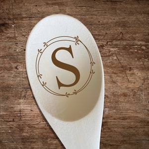 May include: A wooden spoon with a light beige finish. The spoon features a brown letter "S" inside a circular wreath design. The background is a textured, dark brown wooden surface.