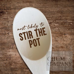 Puede incluir: Cuchara de madera blanca con el texto "most likely to stir the pot" impreso en marrón en la parte delantera.
