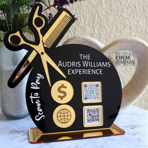 Puede incluir: Premio de acrílico negro y dorado con un par de tijeras y un peine, un signo de dólar, un globo terráqueo y dos códigos QR. El texto "The Audris Williams Experience" y "Chum & Company" están en el premio. El texto "Scan to Pay" está en el lateral del premio.