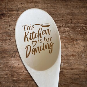 Puede incluir: Una cuchara de madera con el texto "This Kitchen is for Dancing" en marrón. Un pequeño dibujo de sartén está encima de la palabra "Kitchen", y un corazón está entre "Kitchen" y "is". La cuchara está sobre un fondo de madera.