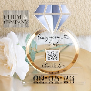 May include: A clear acrylic ring box with a gold band and a diamond-shaped top. The box has a QR code and the text "honeymoon fund" and "Chris & Lisa" printed on it. The box is on a clear acrylic stand with the date "09-05-23" printed on it.