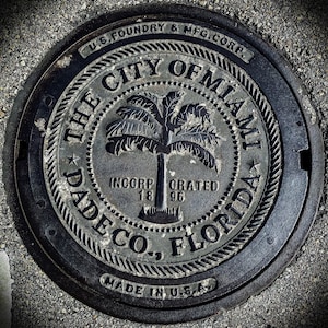 May include: A black metal manhole cover with the text "The City of Miami Dade Co., Florida" and "U.S. Foundry & Mfg. Corp." around the edge. The center of the cover features a palm tree and the text "Incorporated 1896".