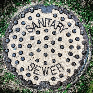 May include: A round, rusty metal sewer cover with a white, sandy surface. The cover has a black, raised inscription that reads "SANITARY SEWER".
