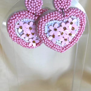 May include: A pair of pink heart-shaped earrings with white and pink flower accents. The earrings are made of beads and sequins.