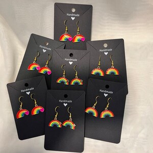 May include: A collection of rainbow earrings with gold hooks, presented on black cards. Each earring showcases a vibrant rainbow design. Some earrings include a heart or star. The cards are labelled "Handmade".