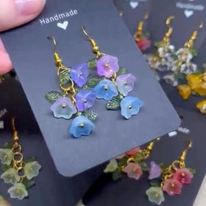 May include: Handmade earrings featuring a cluster of pastel blue, pink, and purple flower charms dangling from gold hoops. The earrings are presented on black cards with the word "Handmade" written on them.