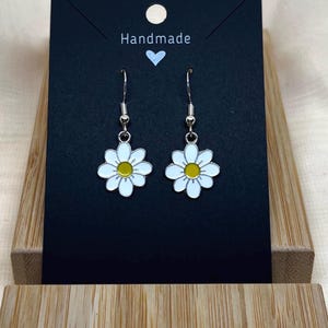 Peut inclure: Une paire de boucles d'oreilles pendantes argentées avec des breloques de marguerite blanches et des centres jaunes. Les boucles d'oreilles sont présentées sur une carte noire avec le mot "Handmade" écrit dessus.