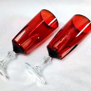 May include: Two red-tinted crystal champagne flutes. The glasses have a faceted design on the bowl and stem, with a clear base. The stems are clear and the base is round. The glasses are angled towards the viewer.