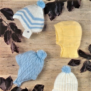 Puede incluir: Cuatro gorros de bebé tejidos en varios estilos y colores. Uno es blanco con rayas azules, otro es azul claro con un diseño de punto de cable, un tercero es amarillo y el último es blanco con rayas verticales. Cada gorro tiene un pompón azul.