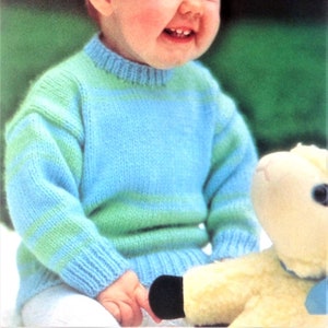 Może przedstawiać: Dziecko w niebiesko-zielonym, prążkowanym swetrze, uśmiecha się i trzyma białą pluszową zabawkę.