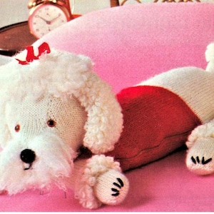 May include: A white knitted poodle dog toy with a red knitted sweater. The toy has a black nose and eyes and is lying on a pink surface.