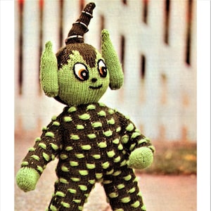 May include: A green knitted alien doll with brown polka dots, large ears, and a pointed head. The doll has orange eyes and a friendly expression.