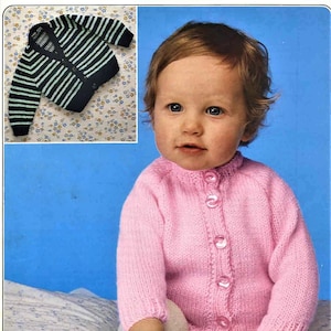 May include: A pink knitted cardigan with button closures, worn by a baby sitting on a white and gray patterned blanket. The baby is wearing white socks and has brown hair.