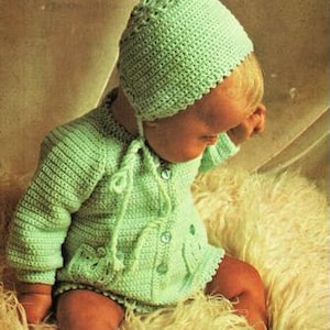 May include: A baby wearing a green crocheted outfit, including a bonnet, cardigan, and booties. The outfit features a delicate floral design.