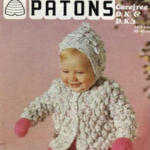 May include: A white knitted baby cardigan with a pink knitted hat and pink knitted booties. The cardigan has a textured pattern and is made with a white yarn with pink flecks. The hat has a pointed top and is decorated with a white flower. The booties are simple and have a pink color. The image includes the text "Patons Carefree D.K. & D.K.'s 18-19 in 46-48 cm".