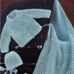 Może przedstawiać: Jasnoniebieski zestaw dla niemowląt z ażurowym splotem, w tym kardigan, dwie czapki i kocyk. Kardigan ma trzy guziki. Czapki mają wstążkę. Kocyk jest prostokątny. Zestaw jest prezentowany na czarnej powierzchni.