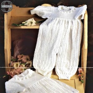 May include: Two white crocheted baby garments, a dress and a one-piece outfit, are displayed on a wooden shelf. Both garments feature a delicate openwork pattern.