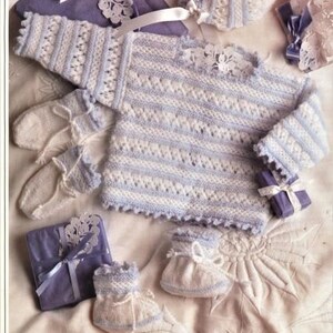 May include: A knitted baby sweater, hat, and booties set in white and light blue with a delicate lace pattern. The set is laid out on a light beige surface with purple gift boxes and lace accents.