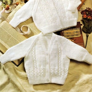 May include: Two white knitted baby cardigans with button fastenings. The cardigan at the top has a round neckline and the cardigan at the bottom has a V-neckline.