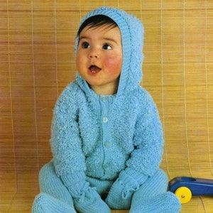 Puede incluir: Un bebé con un mono de punto azul con capucha y cierre de botón. El mono tiene un patrón de punto texturizado y el bebé está sentado en una estera tejida.