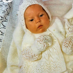 Könnte beinhalten: Ein weißes gestricktes Baby-Outfit mit einem Spitzenmuster. Das Outfit besteht aus einer Strickjacke, einer Mütze und Fäustlingen.
